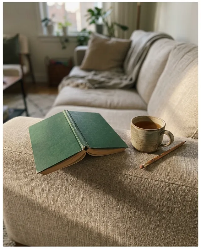 A green paperback face down on the arm of a sofa, next to a mug of tea